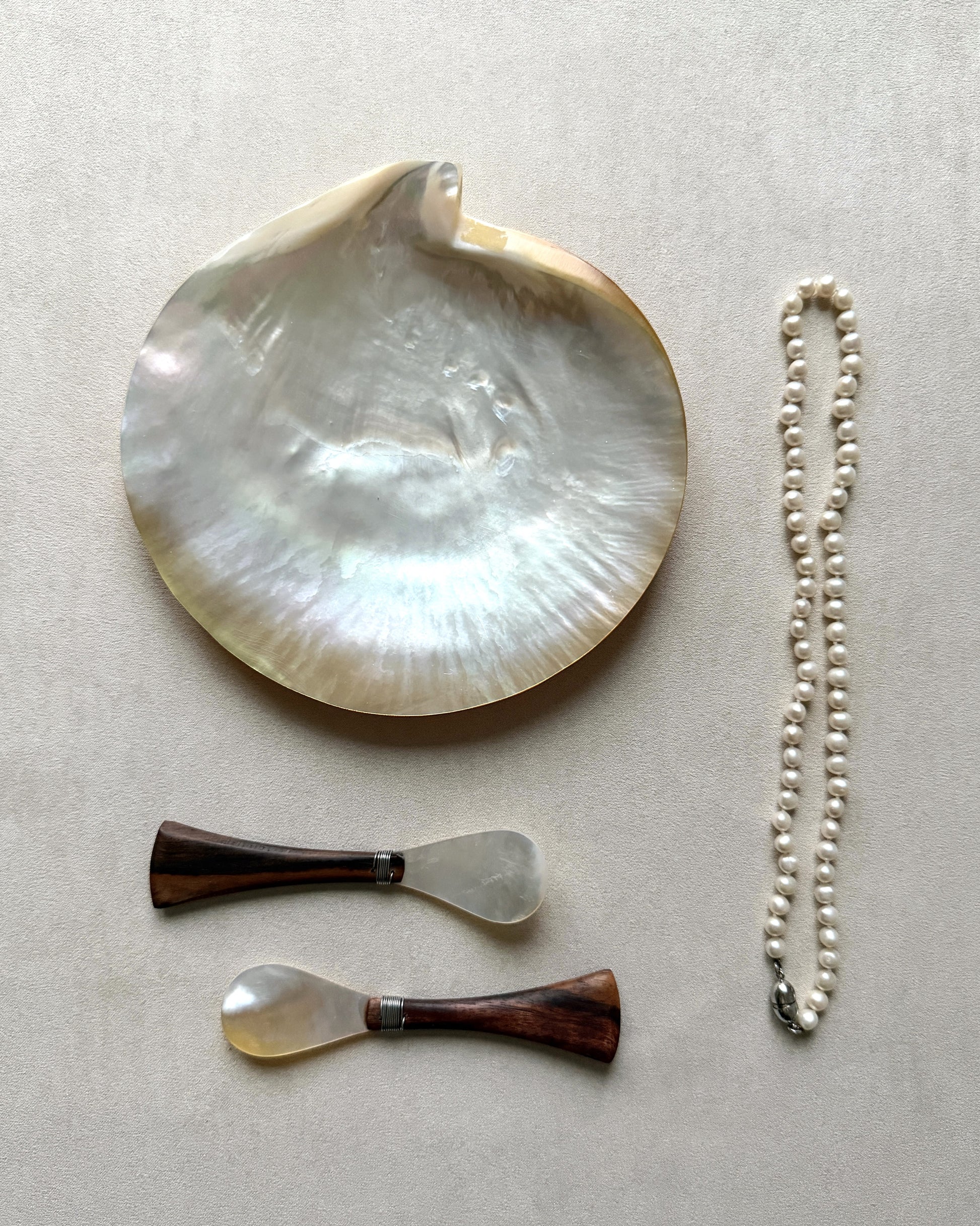 An elegant coastal-inspired flatlay including two teak and pearl spoons, a large natural mother-of-pearl shell, and a pearl necklace on a beige background.