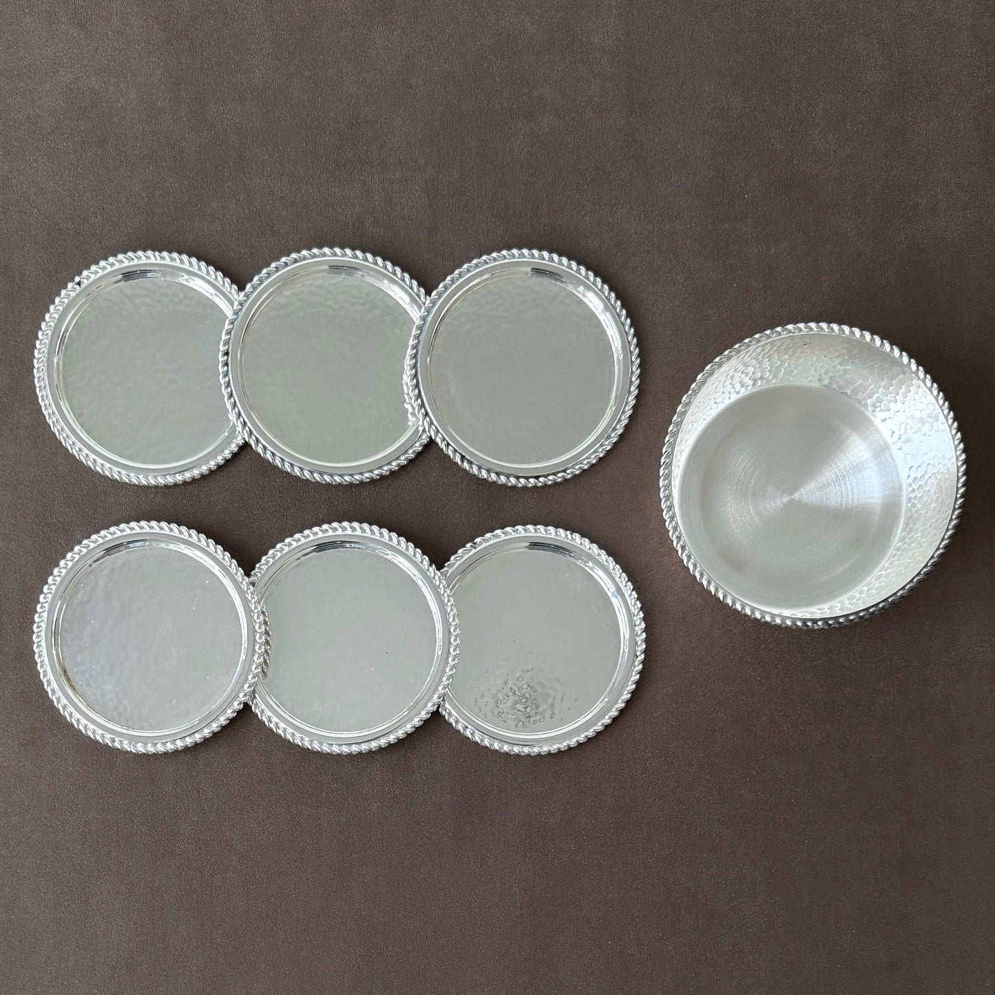 A rare 7-piece hammered silver-plated coaster set from 1960s West Germany, featuring six drink coasters and one wine bottle coaster on a neutral brown background.