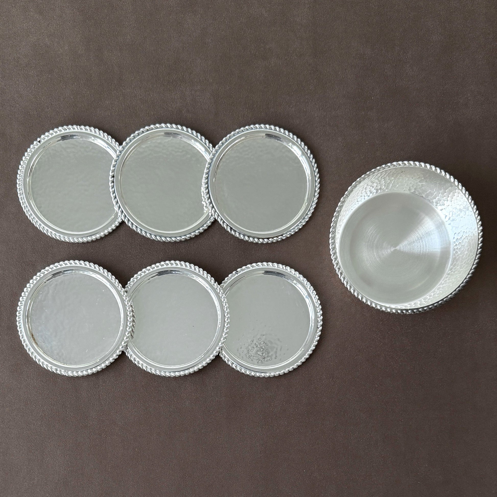 A rare 7-piece hammered silver-plated coaster set from 1960s West Germany, featuring six drink coasters and one wine bottle coaster on a neutral brown background.