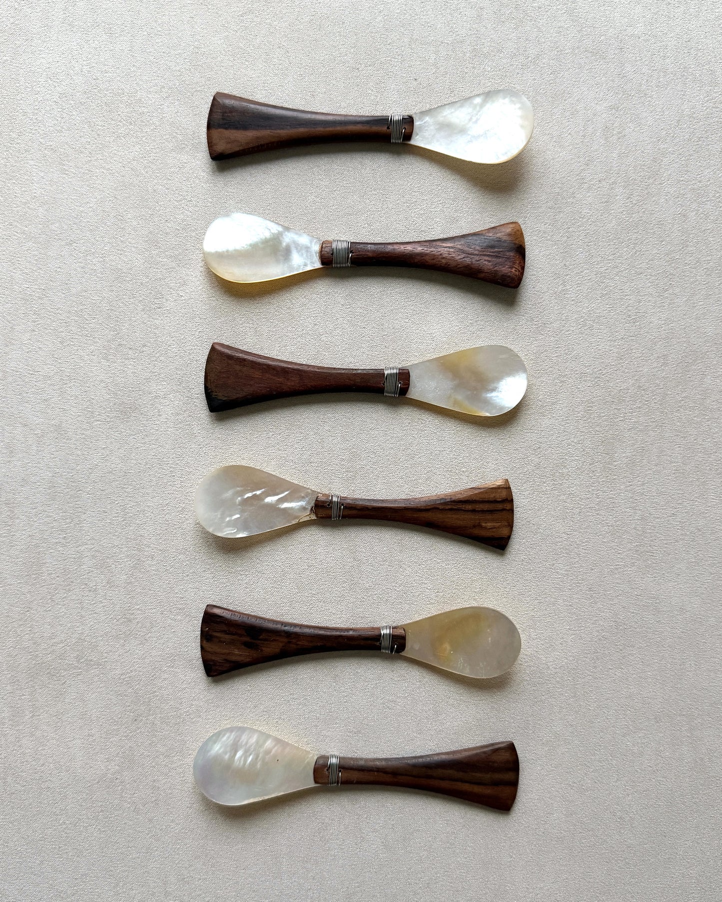 A set of six mid-century modern caviar spoons featuring iridescent mother-of-pearl bowls and tapered teak wood handles on a neutral beige background.
