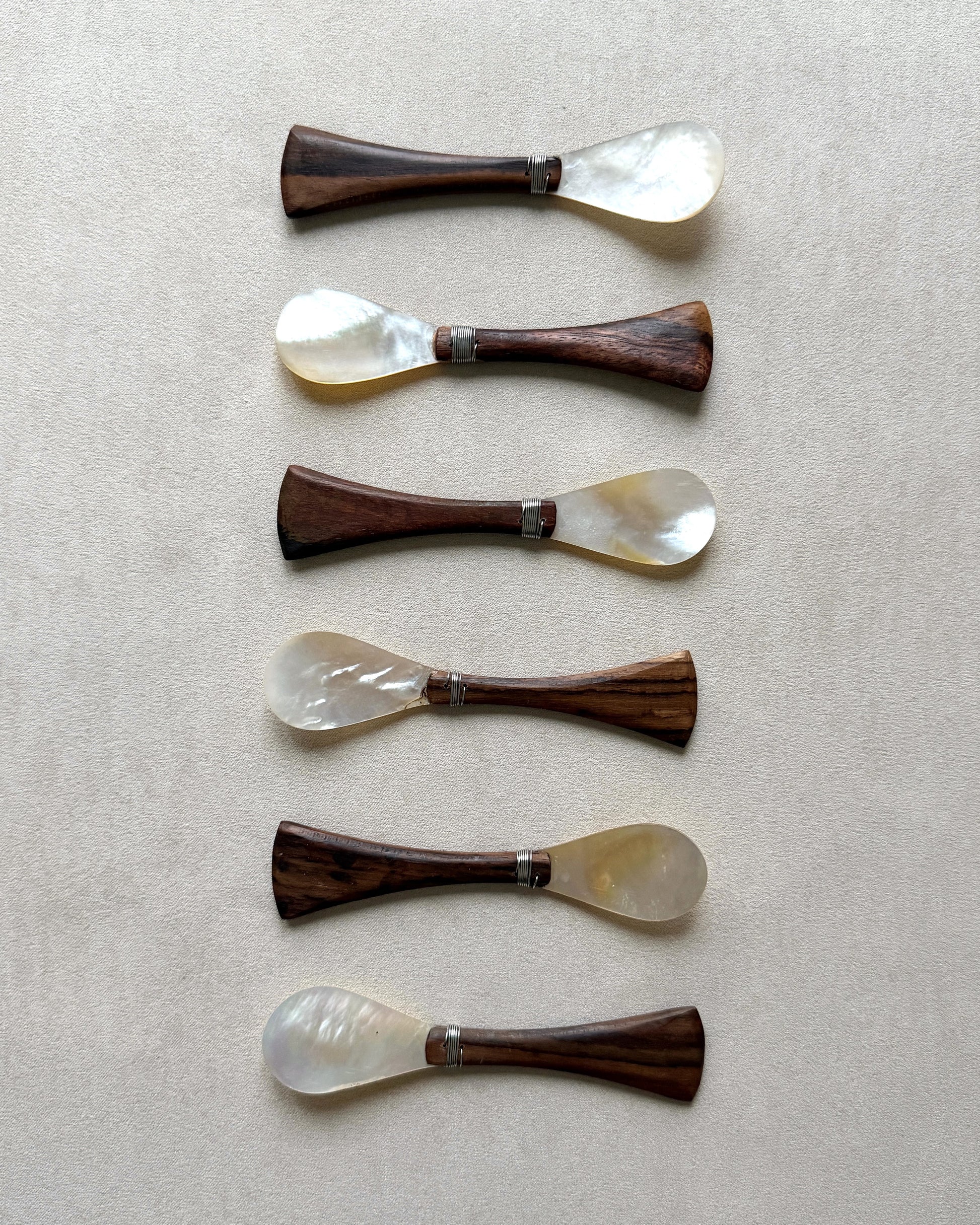 A set of six mid-century modern caviar spoons featuring iridescent mother-of-pearl bowls and tapered teak wood handles on a neutral beige background.