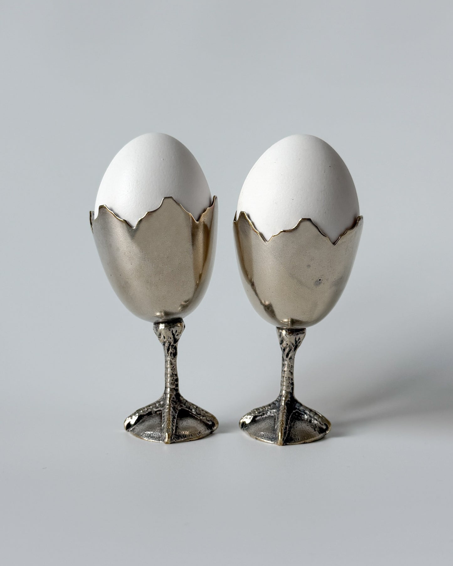 A whimsical 1950s European egg cup made of nickel silver, featuring a sculptural broken eggshell bowl perched on a detailed chicken foot base.