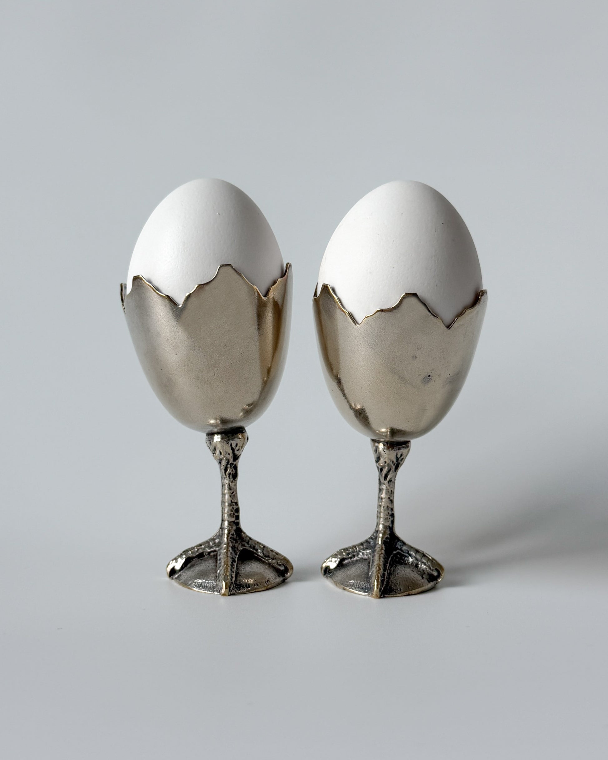 A whimsical 1950s European egg cup made of nickel silver, featuring a sculptural broken eggshell bowl perched on a detailed chicken foot base.