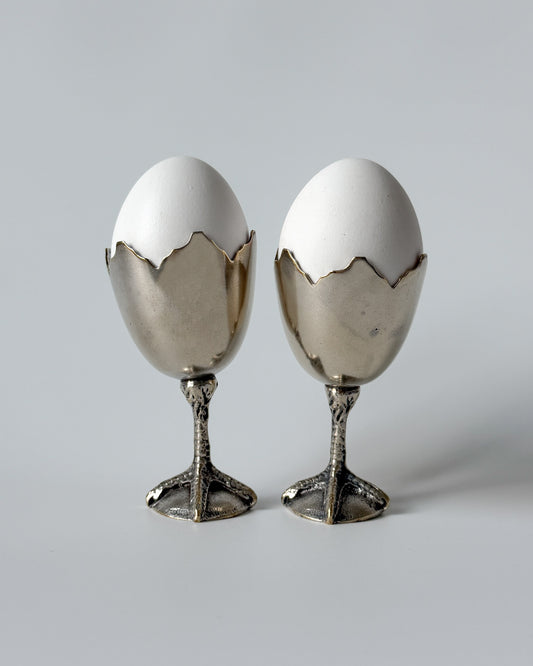 A whimsical 1950s European egg cup made of nickel silver, featuring a sculptural broken eggshell bowl perched on a detailed chicken foot base.