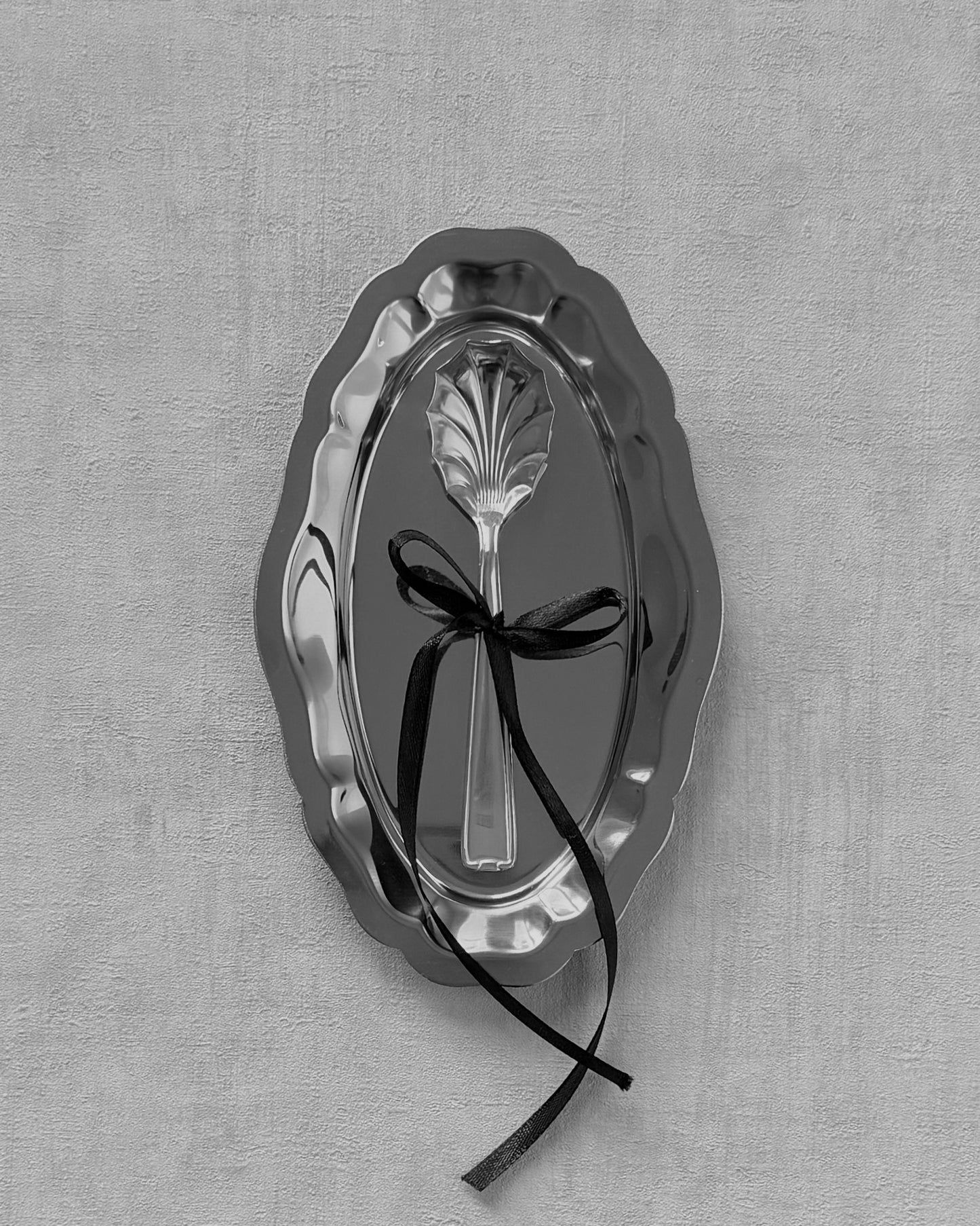 Black and white artistic photograph of a Michelin shell-shaped cream spoon decorated with a black bow on a small vintage silver tray.